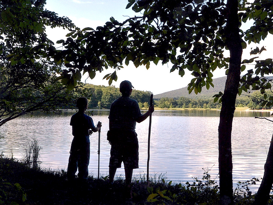 Two young hikers with walking sticks looking out over the lake from behind shade trees
