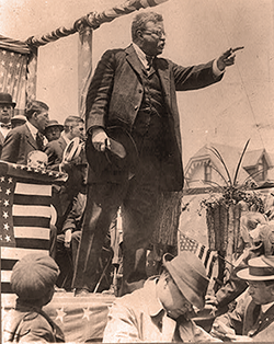 Theodore Roosevelt on a campaign platform, holding his hat in his hand